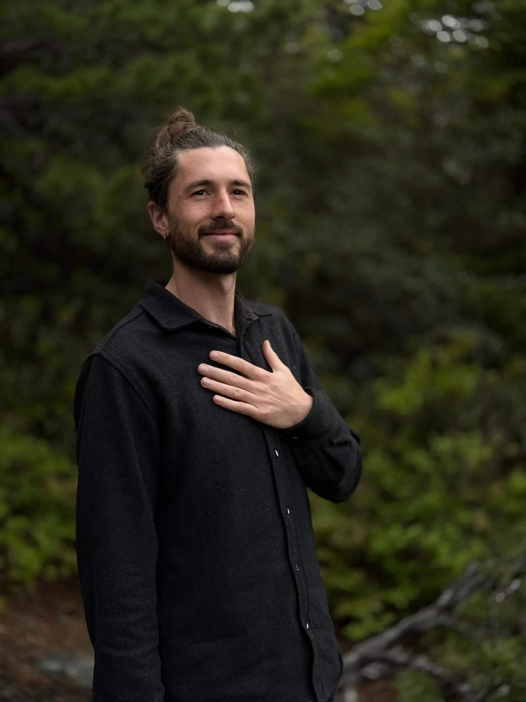 Man in a black shirt with his hand on his heart, standing among evergreen trees