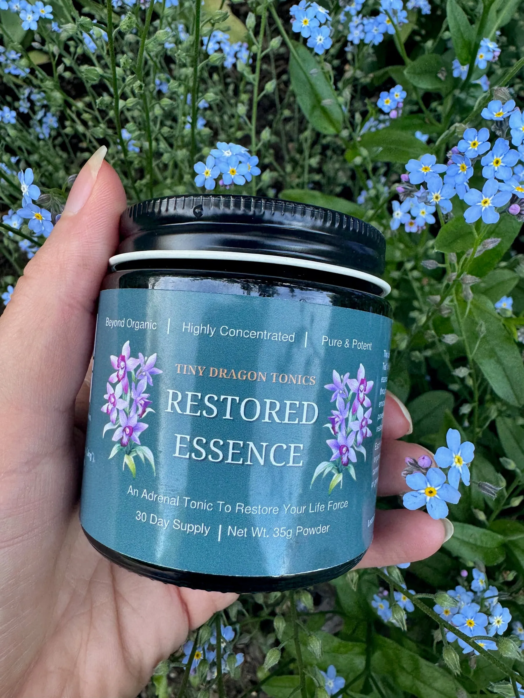 A hand holding a jar of Restored Essence in front of blue forget-me-not flowers